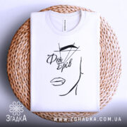 Футболка фея брів, білого кольору, складена на плетеній кошику, фон світлий, принт з малюнком обличчя та написом 'Фея Брів' у чорному кольорі.