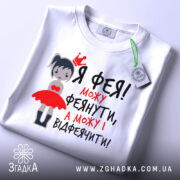 Футболка фея можу феянути, біла футболка на плоскій поверхні, з принтом дівчинки-феї в червоній сукні, текст чорного та червоного кольору, з етикеткою.