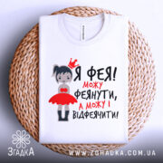 Футболка фея можу феянути, біла футболка на плоскій поверхні з плетеним елементом, з принтом дівчинки-феї в червоній сукні, текст чорного та червоного кольору.