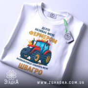 Футболка фермер швагро, біла, зображена зверху, на фоні світло-сірого градієнту, принт з червоним трактором та жовтим текстом, з етикеткою на зеленому шнурку.