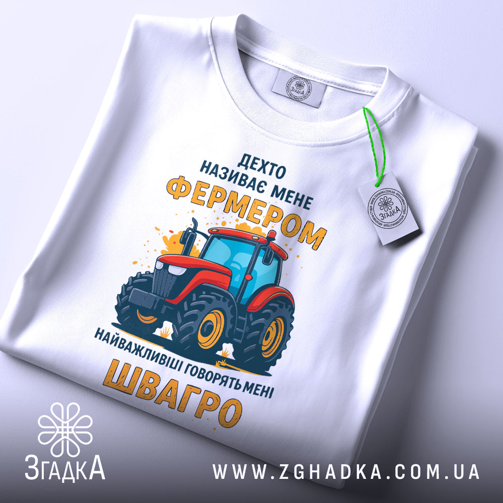 Футболка фермер швагро, біла, зображена зверху, на фоні світло-сірого градієнту, принт з червоним трактором та жовтим текстом, з етикеткою на зеленому шнурку.