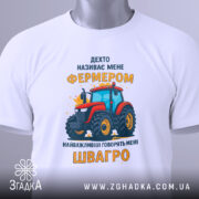 Футболка фермер швагро, біла, зображена зверху, на фоні світло-сірого градієнту, принт з червоним трактором та жовтим текстом, з етикеткою на зеленому шнурку.