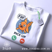 Футболка fih is calling. Біла футболка, складена на поверхні, з принтом рибки, фон світлий, принт чіткий, рибка оранжевого кольору з великими очима, текст 'FiH' та 'PICK UP' під рибкою, на футболці є етикетка з зеленим шнурком.