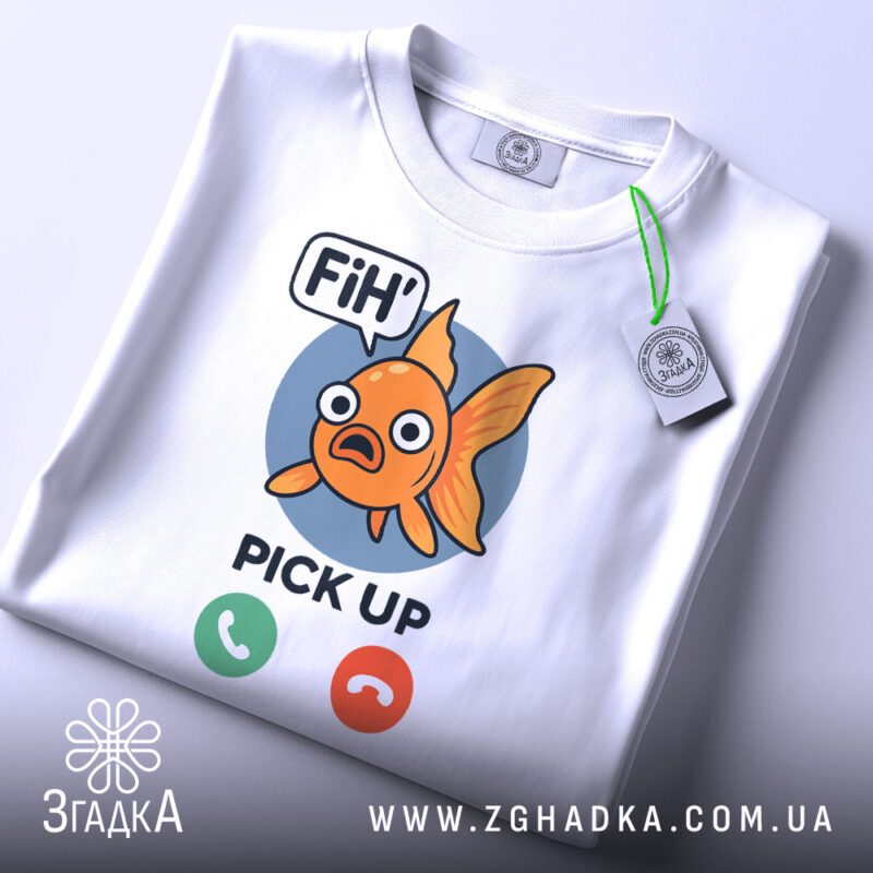 Футболка fih is calling. Біла футболка, складена на поверхні, з принтом рибки, фон світлий, принт чіткий, рибка оранжевого кольору з великими очима, текст 'FiH' та 'PICK UP' під рибкою, на футболці є етикетка з зеленим шнурком.