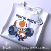 Футболка фітнес настрій, біла, складена на столі, фон світло-сірий, принт з персонажем, що піднімає штангу, текст 'TRUST THE PROCESS' над ним, з етикеткою на рукаві.