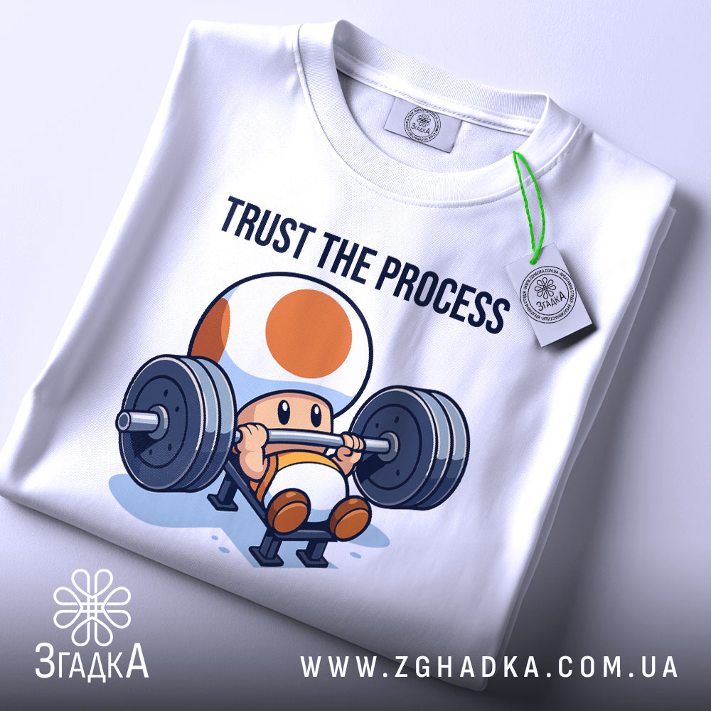Футболка фітнес настрій, біла, складена на столі, фон світло-сірий, принт з персонажем, що піднімає штангу, текст 'TRUST THE PROCESS' над ним, з етикеткою на рукаві.