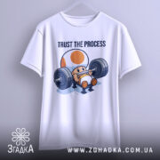 Футболка фітнес настрій, біла, підвішена на вішалці, фон світло-сірий, принт з персонажем, що піднімає штангу, текст 'TRUST THE PROCESS' над ним.