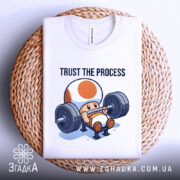 Футболка фітнес настрій, біла, складена на плетеній основі, фон світло-сірий, принт з персонажем, що піднімає штангу, текст 'TRUST THE PROCESS' над ним.