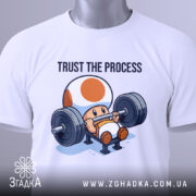 Футболка фітнес настрій, біла, складена на столі, фон світло-сірий, принт з персонажем, що піднімає штангу, текст 'TRUST THE PROCESS' над ним, з етикеткою на рукаві.