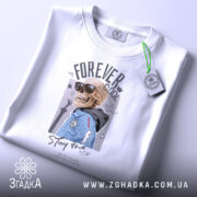 Футболка forever cool, білого кольору, складена на столі, фон світло-сірий, принт з черепом у сонцезахисних окулярах та синій куртці, на комірці видна етикетка.