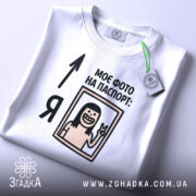 Футболка фото на паспорт: складена біла футболка з принтом, на якому зображена усміхнена людина з довгим волоссям, що показує знак миру, текст "МОЄ ФОТО НА ПАСПОРТ:" розміщений зверху, на футболці є етикетка з зеленим шнурком, фон світло-сірий.