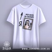 Футболка фото на паспорт: біла футболка, підвішена на вішалці, з принтом, що містить малюнок усміхненої людини з довгим волоссям, яка показує знак миру, текст "МОЄ ФОТО НА ПАСПОРТ:" розташований зверху, фон світло-сірий.