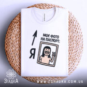 Футболка фото на паспорт: складена біла футболка на плетеній кошику, з принтом, що містить малюнок усміхненої людини з довгим волоссям, яка показує знак миру, текст "МОЄ ФОТО НА ПАСПОРТ:" розміщений зверху, фон білий.