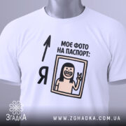 Футболка фото на паспорт: складена біла футболка з принтом, на якому зображена усміхнена людина з довгим волоссям, що показує знак миру, текст "МОЄ ФОТО НА ПАСПОРТ:" розміщений зверху, на футболці є етикетка з зеленим шнурком, фон світло-сірий.