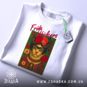 Футболка Фріда стиль, біла, складена на столі, фон світло-сірий, принт з котом у стилі Фріди Кало, з червоними квітами та написом 'Frida Whiskers', на етикетці зелена стрічка.