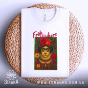 Футболка Фріда стиль, біла, складена на плетеному кошику, фон світло-сірий, принт з котом у стилі Фріди Кало, з червоними квітами та написом 'Frida Whiskers'.