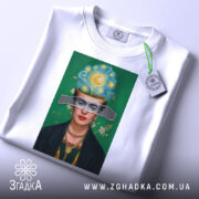 Футболка Фріда білого кольору, складена на столі, фон світлий, принт з портретом жінки з яскравим головним убором, на ярлику зелена стрічка.