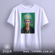 Футболка Фріда білого кольору, підвішена на вішалці, фон сірий, принт з портретом жінки з яскравим головним убором та зеленим фоном.