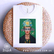 Футболка Фріда білого кольору, складена на плетеній кошику, фон світлий, принт з портретом жінки з яскравим головним убором, видима етикетка.