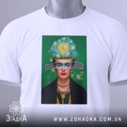 Футболка Фріда білого кольору, складена на столі, фон світлий, принт з портретом жінки з яскравим головним убором, на ярлику зелена стрічка.