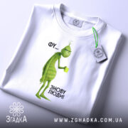 Футболка фу люди. Біла футболка, складена на поверхні, з принтом зеленого персонажа, що тримає чашку. Фон світлий, ракурс зверху, видно етикетку з логотипом, текст на принті: 'ФУ... ЗНОВУ ЛЮДИ!'