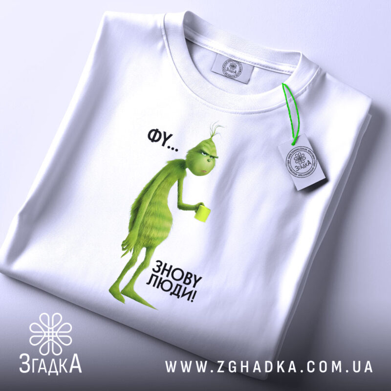 Футболка фу люди. Біла футболка, складена на поверхні, з принтом зеленого персонажа, що тримає чашку. Фон світлий, ракурс зверху, видно етикетку з логотипом, текст на принті: 'ФУ... ЗНОВУ ЛЮДИ!'