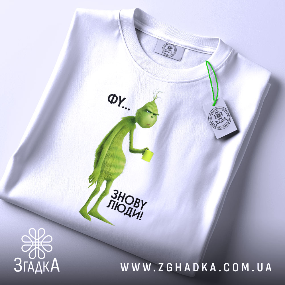 Футболка фу люди. Біла футболка, складена на поверхні, з принтом зеленого персонажа, що тримає чашку. Фон світлий, ракурс зверху, видно етикетку з логотипом, текст на принті: 'ФУ... ЗНОВУ ЛЮДИ!'