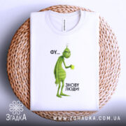 Футболка фу люди. Біла футболка, складена на плетеній основі, з принтом зеленого персонажа з чашкою. Фон світлий, ракурс зверху, текст на принті: 'ФУ... ЗНОВУ ЛЮДИ!'