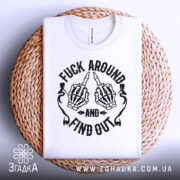 Футболка fuck around find out, білого кольору, складена на плетеній кошику, з верхнього ракурсу, фон світло-сірий, принт з чорними скелетними руками, що показують середній палець, з написом 'AND' та 'FIND OUT'.