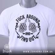 Футболка fuck around find out, білого кольору, складена на поверхні, з верхнього ракурсу, фон світло-сірий, принт з чорними скелетними руками, що показують середній палець, з написом 'AND' та 'FIND OUT', на етикетці зелена стрічка.