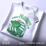 Футболка Glorbo Fruttodrillo білого кольору, зображена в горизонтальному положенні, фон світлий, принт з зеленим крокодилом, що сидить на воді, з елементами природи, на комірці видно етикетку з зеленим шнурком.