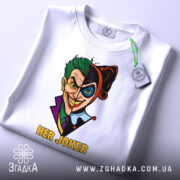 Футболка гарлі джокер білого кольору, розкладена на поверхні, з принтом, що зображає Джокера та Харлі Квін, з текстом 'HER JOKER', на футболці є етикетка з зеленим шнурком, фон світло-сірий.