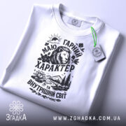 Футболка гарний характер, біла, складена на столі, фон світло-сірий, принт чорного кольору з зображенням лева, гір, річки та квітів, з етикеткою зеленого кольору.