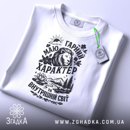 Футболка гарний характер, біла, складена на столі, фон світло-сірий, принт чорного кольору з зображенням лева, гір, річки та квітів, з етикеткою зеленого кольору.