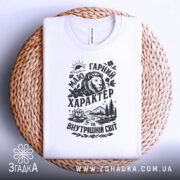 Футболка гарний характер, біла, складена на плетеному кошику, фон білий, принт чорного кольору з зображенням лева, гір, річки та квітів.