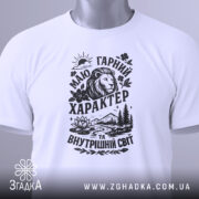Футболка гарний характер, біла, складена на столі, фон світло-сірий, принт чорного кольору з зображенням лева, гір, річки та квітів, з етикеткою зеленого кольору.