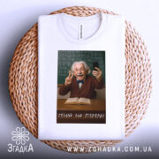 Футболка геній на перерві. Біла футболка, на якій зображений усміхнений чоловік з сивим волоссям, що тримає телефон і показує знак миру. Футболка лежить на плетеній основі, а фон складається з зеленої дошки з формулами. Текст на футболці: 'ГЕНІЙ НА ПЕРЕРВІ'.