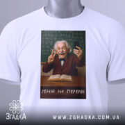 Футболка геній на перерві. Біла футболка, на якій зображений усміхнений чоловік з сивим волоссям, що тримає телефон і показує знак миру. Принт розташований на грудях, а на фоні видно зелену дошку з формулами. Текст на футболці: 'ГЕНІЙ НА ПЕРЕРВІ'.