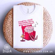 Футболка генії потребують відпочинку, біла, зображена зверху, на фоні плетеного кошика, принт з малюнком мозку в окулярах, що сидить у кріслі, текст червоним шрифтом.