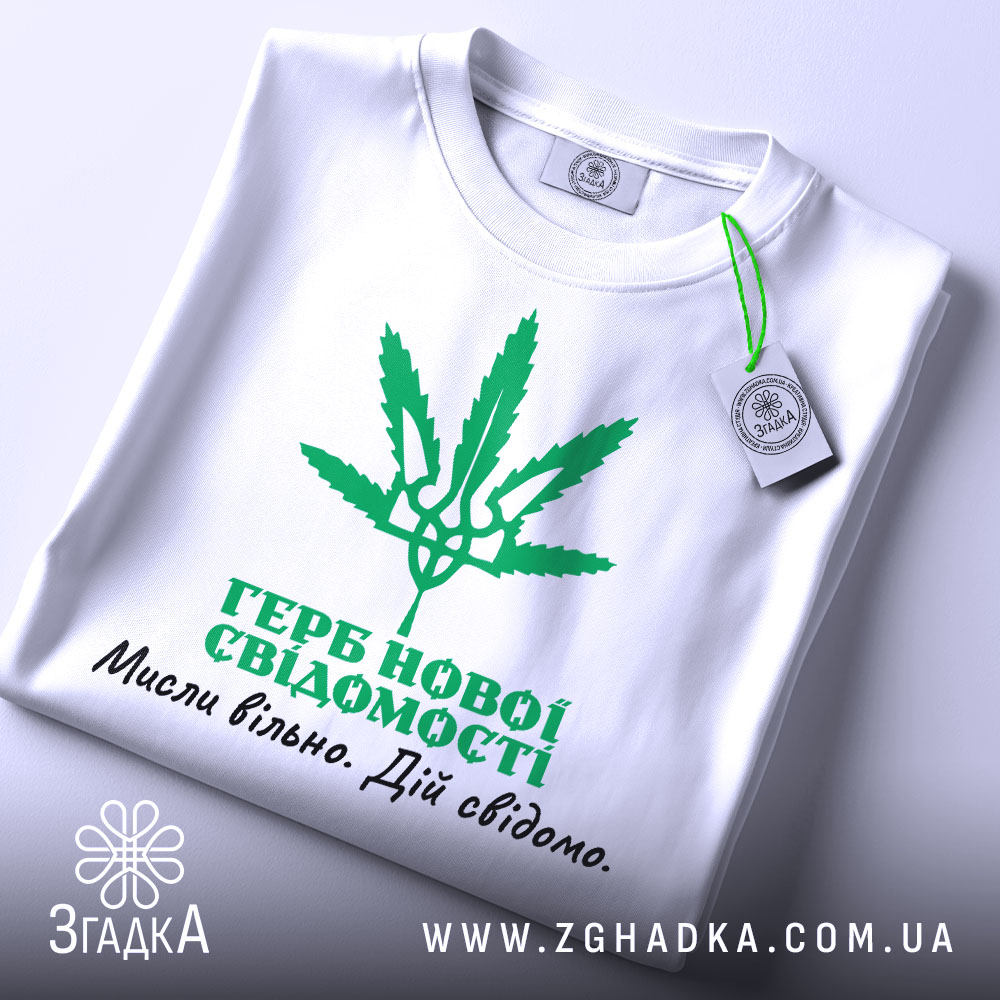 Футболка герб нової свідомості, біла, зображена зверху, складена на столі, фон світло-сірий, принт зелений з зображенням листа коноплі та текстом українською мовою, на комірці є етикетка.