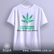 Футболка герб нової свідомості, біла, зображена на вішалці, фон світло-сірий, принт зелений з зображенням листа коноплі та текстом українською мовою.