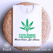 Футболка герб нової свідомості, біла, зображена складеною на плетеному кошику, фон світло-сірий, принт зелений з зображенням листа коноплі та текстом українською мовою.