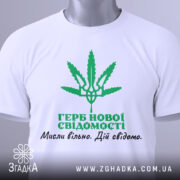 Футболка герб нової свідомості, біла, зображена зверху, складена на столі, фон світло-сірий, принт зелений з зображенням листа коноплі та текстом українською мовою, на комірці є етикетка.