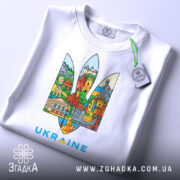 Футболка герб Україна, біла, складена на столі, фон світло-сірий, принт містить кольорові зображення українських архітектурних пам'яток у формі тризуба, текст 'UKRAINE' виконаний у блакитному кольорі, на футболці є етикетка з зеленим шнурком.