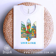 Футболка герб Україна, біла, складена на плетеній кошику, фон світло-сірий, принт містить кольорові зображення українських архітектурних пам'яток у формі тризуба, текст 'UKRAINE' виконаний у блакитному кольорі.