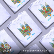 Футболка герб Україна, біла, зображена на манекені, фон світло-сірий, принт містить кольорові зображення українських архітектурних пам'яток у формі тризуба, текст 'UKRAINE' виконаний у блакитному кольорі.
