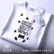 Футболка герой татко, біла, складена на поверхні, фон світло-сірий, принт чорного кольору з текстом "Навіщо потрібні супер-герої, якщо є татко" та малюнком супергероя, на футболці є етикетка з зеленим шнурком.