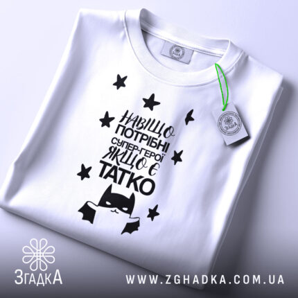 Футболка герой татко, біла, складена на поверхні, фон світло-сірий, принт чорного кольору з текстом "Навіщо потрібні супер-герої, якщо є татко" та малюнком супергероя, на футболці є етикетка з зеленим шнурком.
