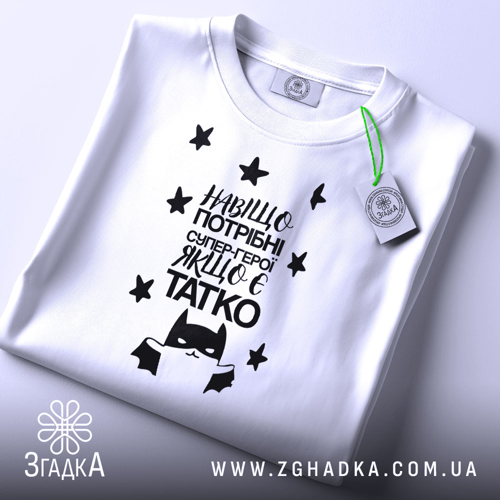 Футболка герой татко, біла, складена на поверхні, фон світло-сірий, принт чорного кольору з текстом "Навіщо потрібні супер-герої, якщо є татко" та малюнком супергероя, на футболці є етикетка з зеленим шнурком.