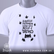 Футболка герой татко, біла, складена на поверхні, фон світло-сірий, принт чорного кольору з текстом "Навіщо потрібні супер-герої, якщо є татко" та малюнком супергероя, на футболці є етикетка з зеленим шнурком.